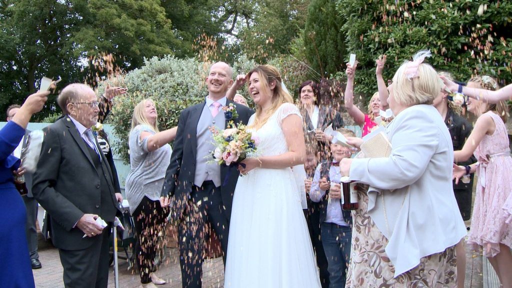 guests laugh and shout out as they shower the bride and groom in dried rose wedding confetti for the wedding video at Abels Harp