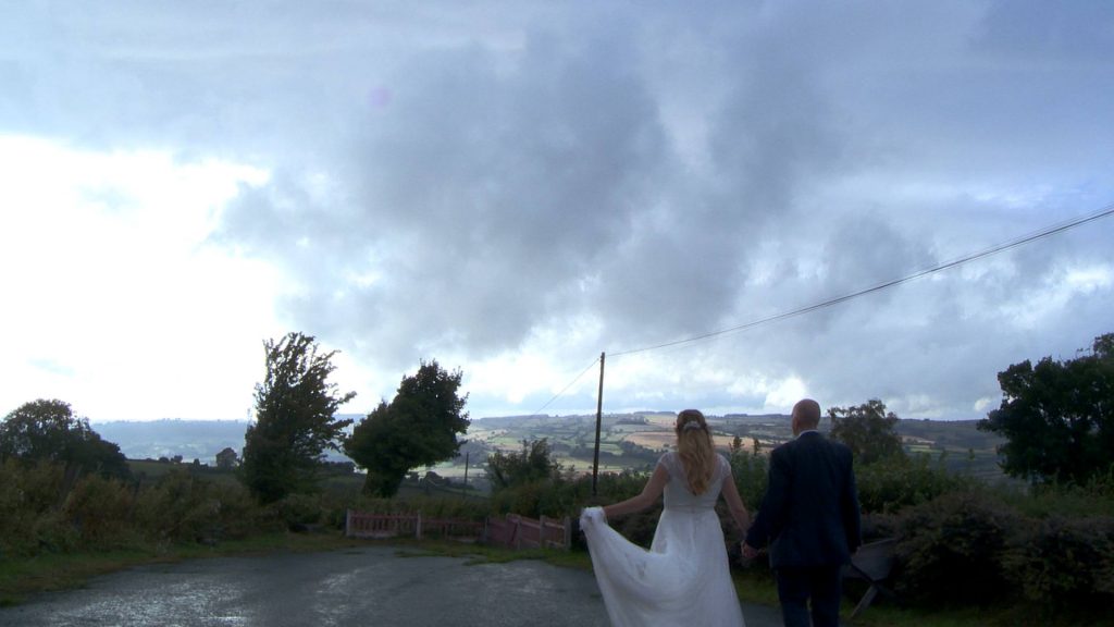 the bride holds her dress up as the married couple go for a walk towards the welsh hills. dark, dramatic clouds hover above them