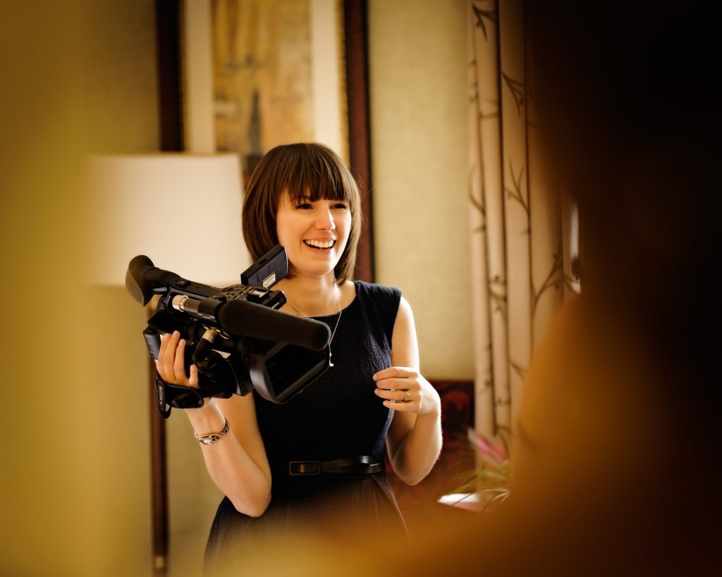 Charlene from Love Gets Sweeter a female wedding videographer laughing whilst holding a Sony video camera in a bridal suite at Mottram Hall in Cheshire