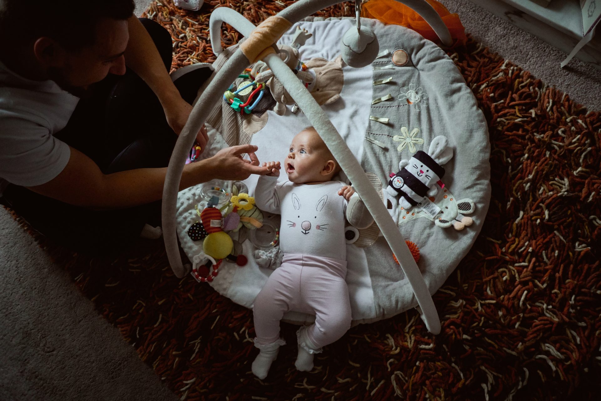 baby girl Freiya lays on her grey and cream play mat holding her daddys finger tightly and smiling up at him during a family newborn photography shoot with love gets sweeter in Lancashire