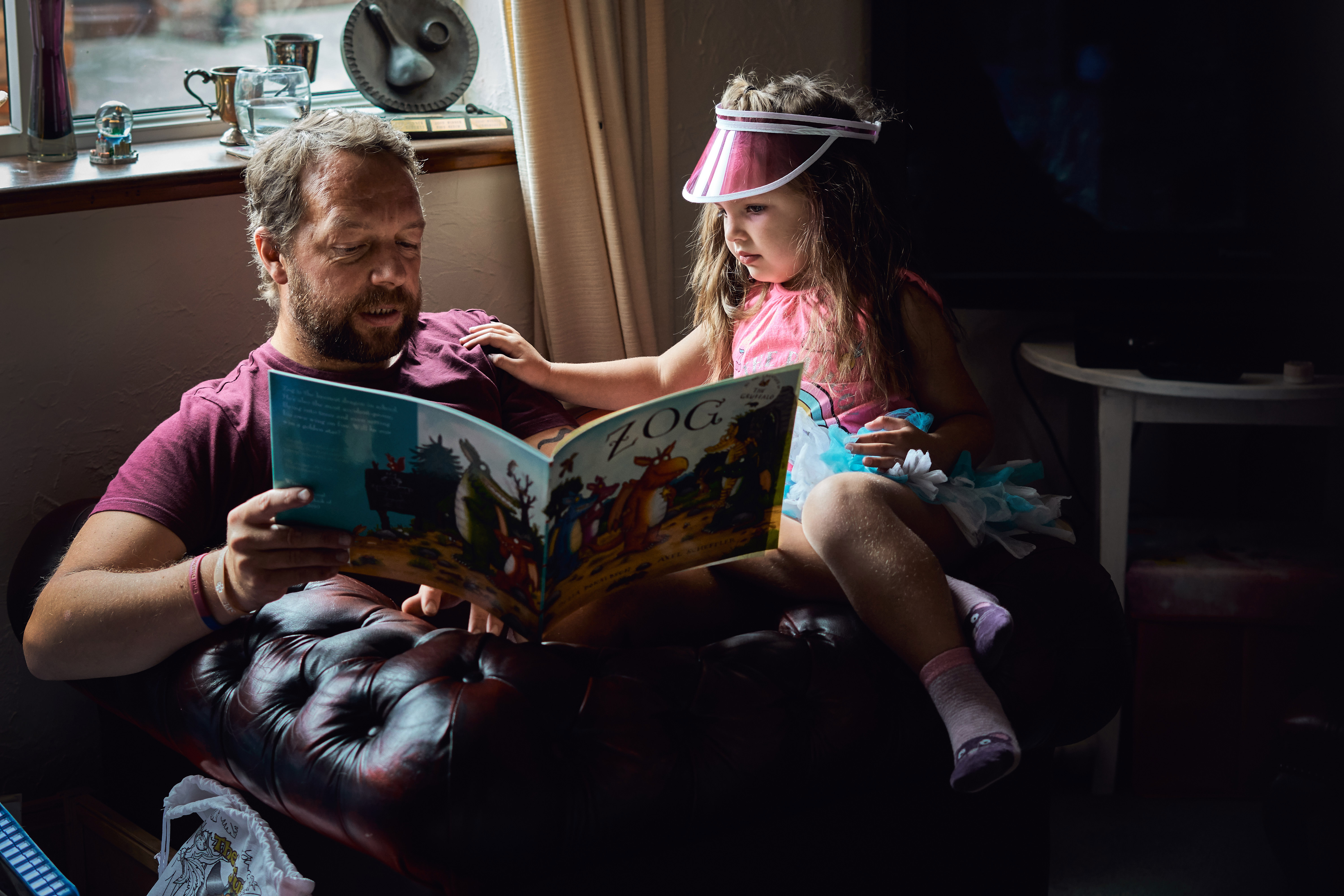 a daughter sits with her dad in a deep red leather armchair in front of a window reading the book Zog together. She's wearing a fun pink visor hat and they're just chilling during a relaxed family photo and video shoot at their home in Newburgh Lancashire