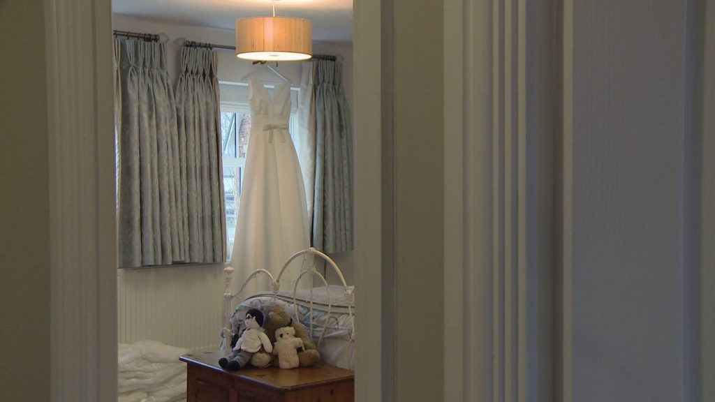 A beautiful simple and elegant silk wedding dress hangs on the curtain pole in a bedroom at the brides parents home in Preston