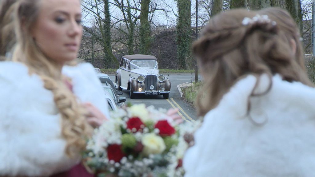 we can see a wedding car turning down the lane towards Broughton Church with the waiting bridesmaids wearing red and white