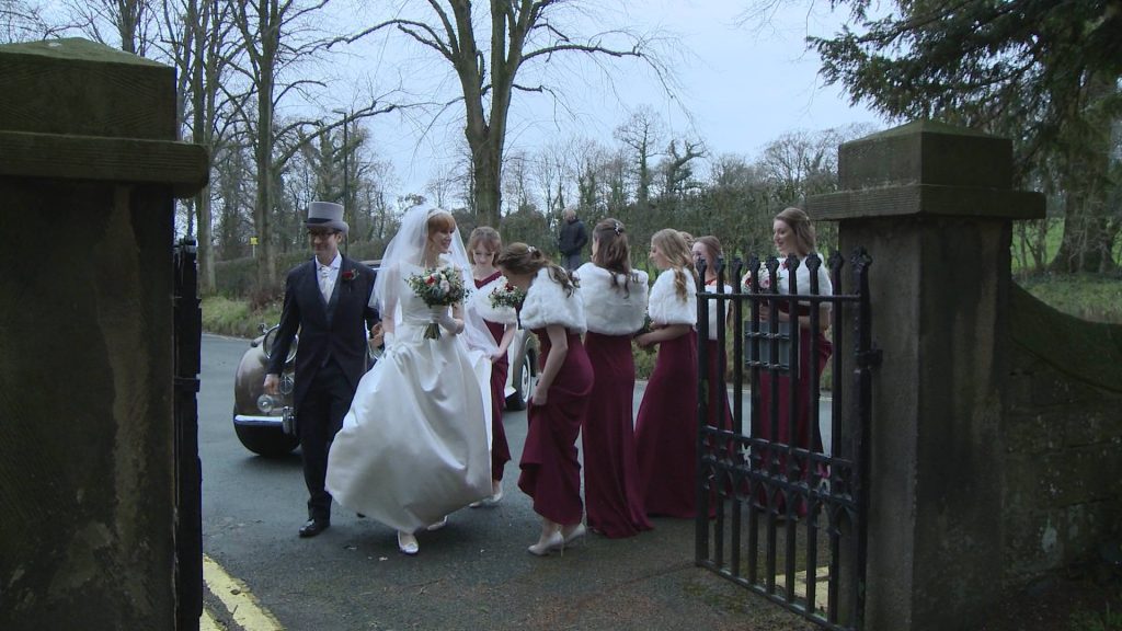 a shot from the wedding video of the dad and bride walking towards the church along with the bridesmaids wearing red full length dresses and fur shrugs capes outside Broughton Church