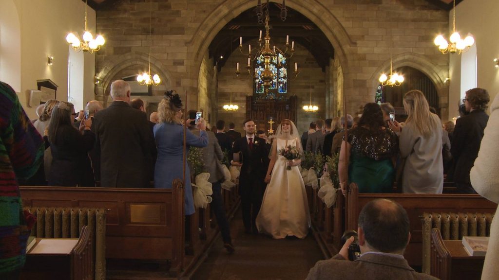 the bride and groom smile and walk back down the aisle at Broughton St Johns Church towards the wedding videographer