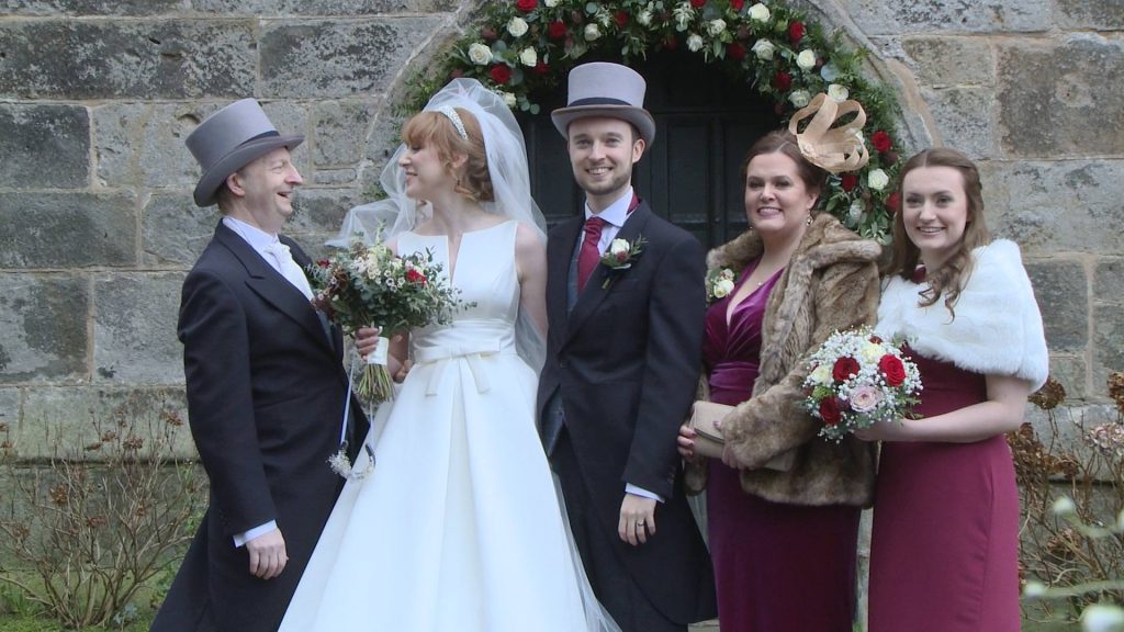 the bride laughs with her new father in law as he catches the wedding veil outside the historical doors at Broughton Church