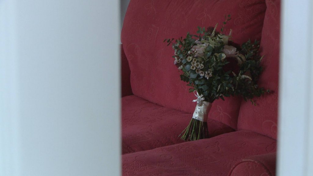 a rustic deep red and pink hand tied wedding bouquet sits on a faded red sofa. the wedding videographer is filming it through a gap in the door at a home in Preston