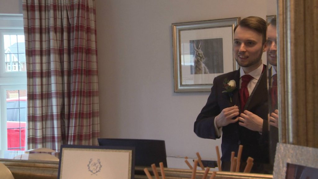 the groom stands in front of a mirror as the wedding videographer films him doing his last wedding suit checks before they leave for the church in Broughton
