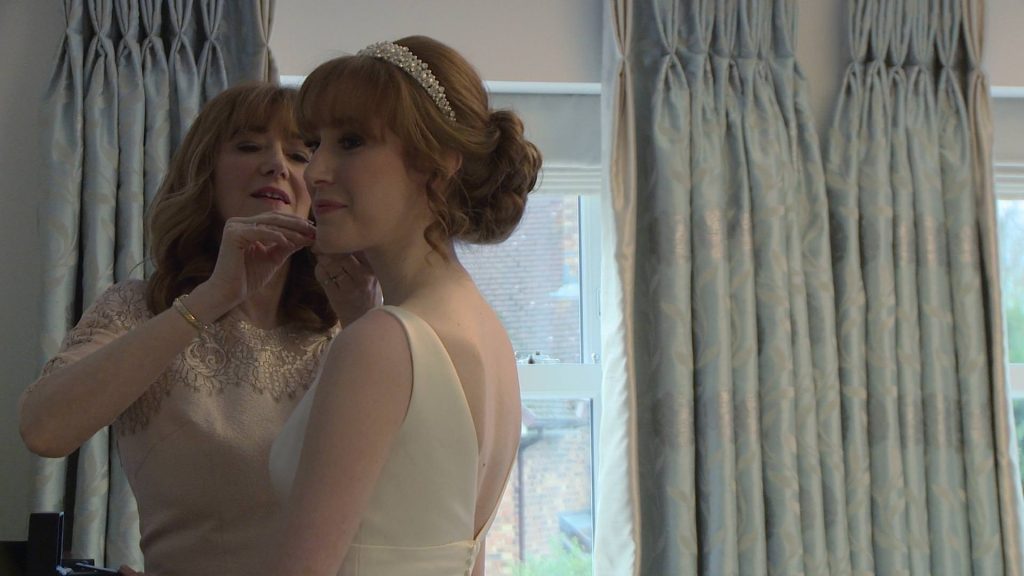 a mother of the bride helps her daughter the bride put her wedding earrings in in her bedroom . she stands so happy and elegant as the videographer films her getting ready in Preston