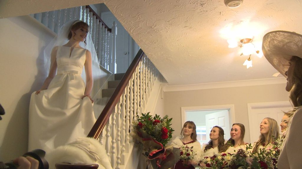 a bride walks down the stairs in her parents home to her awaiting bridesmaids as they gasp and cry seeing her. Caught for their wedding video by wedding videographer love gets sweeter