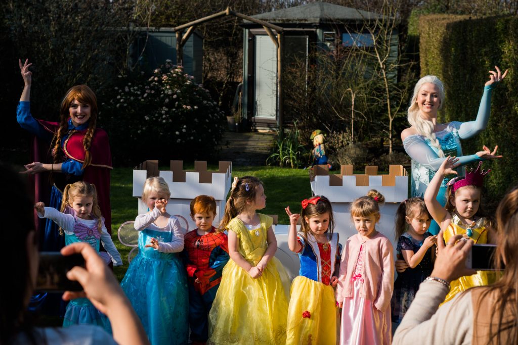a group of 4 year olds dressed as disney princesses line up to pose in front of a cardboard castle in a family garden alongside Elsa and Anna from enchanted encounters in the north west. A natural documentary style professional photograph by Love Gets Sweeter in Burscough