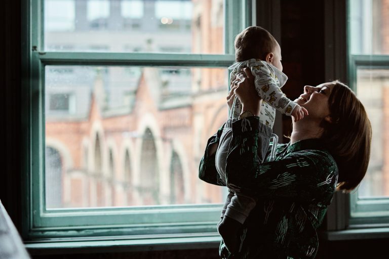 a mum lifts her baby boy up and smiles in front of a window for a family photography workshop in Manchester