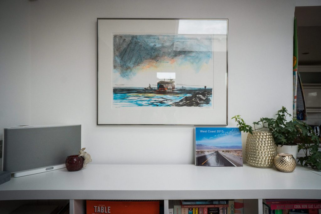 A wide shot of a kitchen wall with a beautiful painting of a boat hanging up and a family holiday photobook next to a candle holder and plant pot and sonos speaker