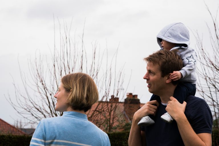 a little girl sits on her dads shoulders and holds on to his ears during a fun family shoot in their garden near Manchester