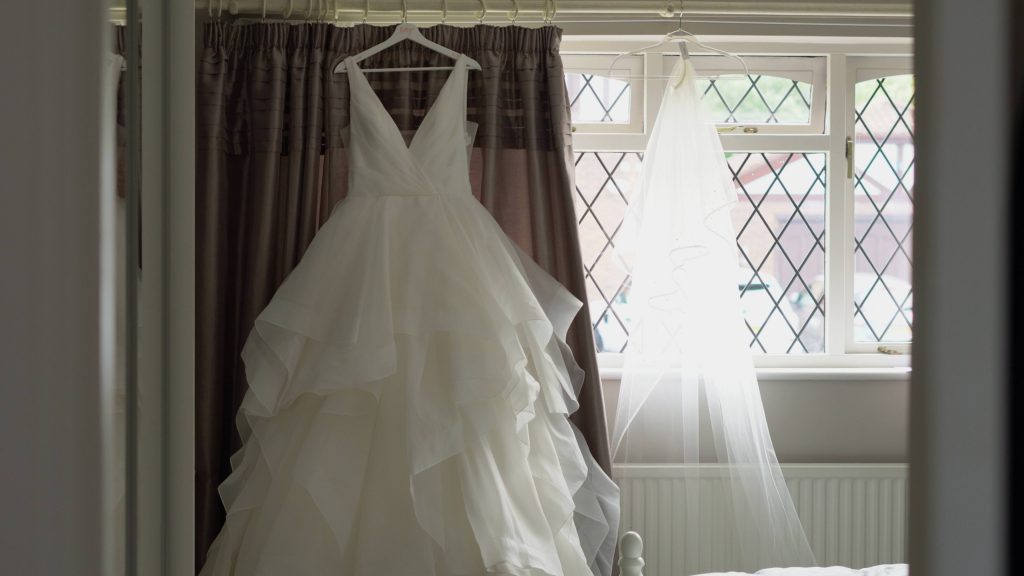 a white princess style tulle layered wedding dress hangs on a curtain rail next to the brides veil ready for the bride getting married in the wrral