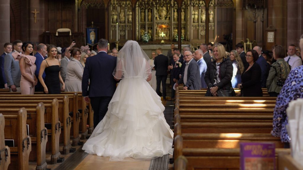 A wedding video still of a bride walking down the aisle in a princess style wedding dress at Oxton St Saviours in the wirral