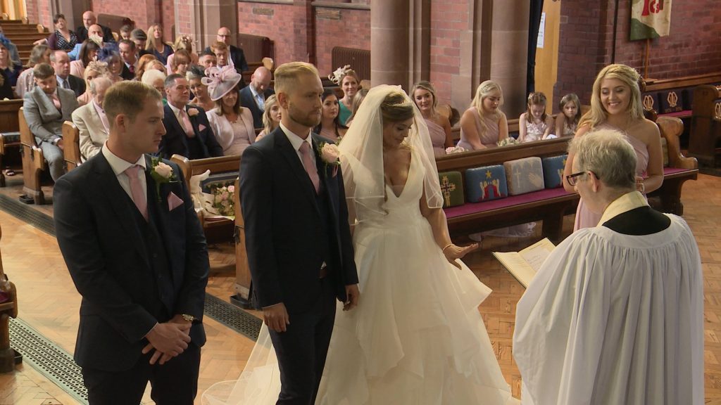 a wide shot from the wedding videographer as the bride looks down at her new wedding ring as she holds hands with her new husband at a church wedding in the wirral