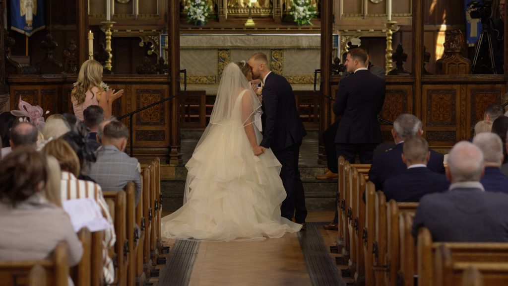 a wide shot from the wedding video at the top of the aisle as the bride and groom have their first kiss at oxton st saviours church
