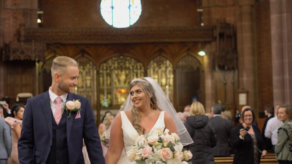 a still from the wedding video of a bride and groom walking down the aisle now married at oxton st saviours