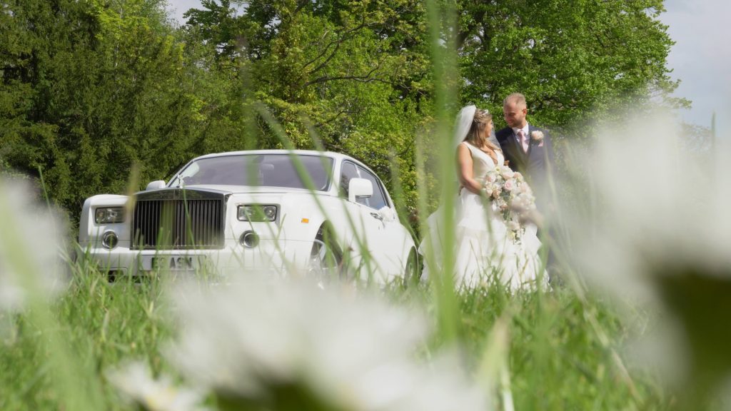 a still from the wedding videographer of the white rolls royce phantom wedding car next to the bride and groom in the gardens at Thornton Manor