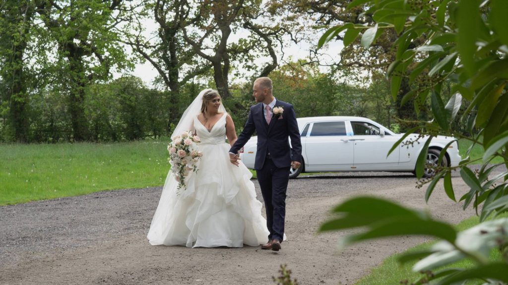 a bride wearing a princess wedding dress and groom in navy walk away from their white rolls royce wedding car at thornton manor