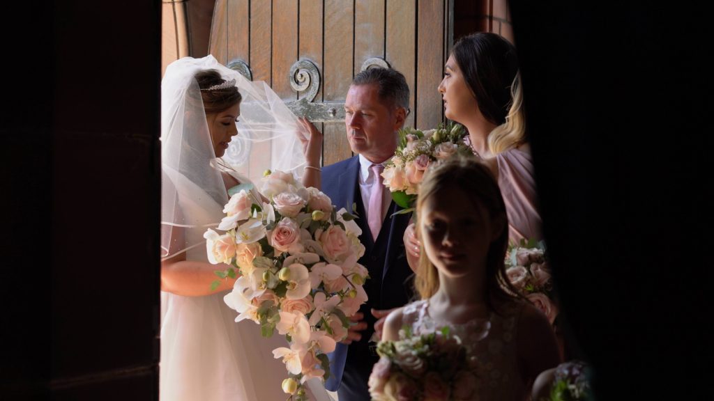 a beautiful still from the wedding videographer of the bride having her veil put in place by her dad moments before she walks down the aisle at Oxton St Saviours