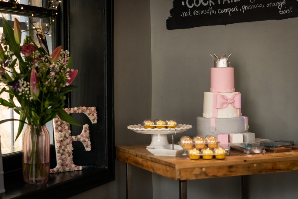 a 3 tier pink, white and grey christening cake with a bow and princess crown on sits on a table surrounded by cupcakes and a giant F for Florence in Site pizzeria ahead of a christening party