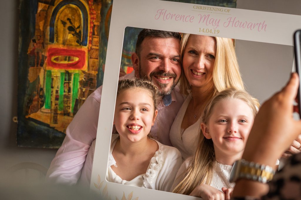 a family photographer gets a photo of someone taking a photo during a christening party of a family of four holding an Instagram frame