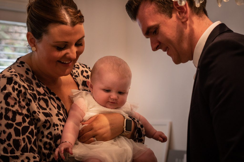 a photograph of a mum holds her baby girl in her christening outfit and smiles as dad tries to get her little shoes on ready for her christening