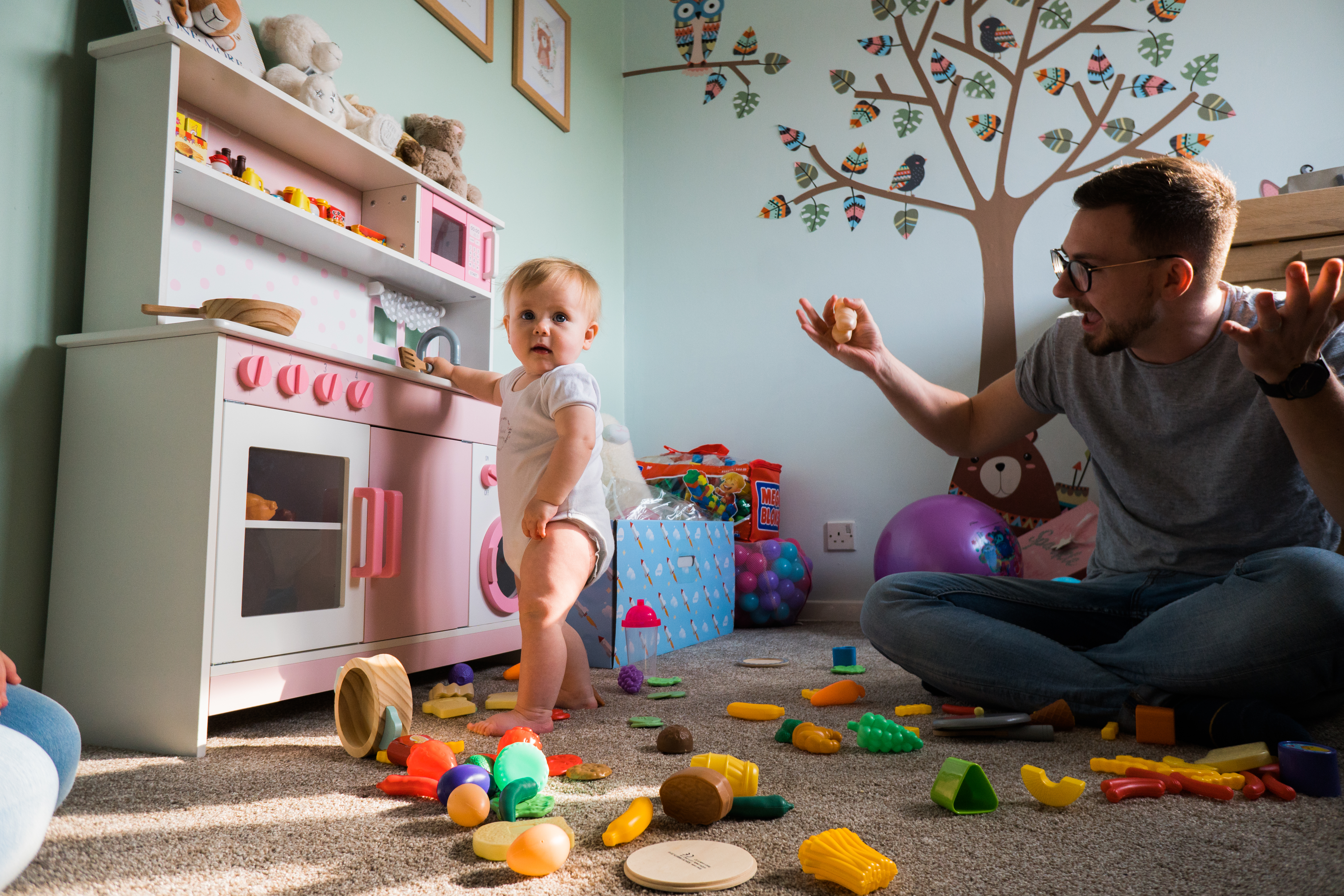 a dad sits in a nursery with his arms up at the mes his daughter has made with her play kitchen