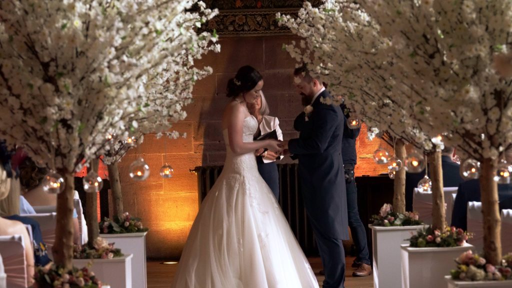a wide shot from the wedding video of an aisle of white blossom trees at a wedding cermony at Peckforton castle in cheshire
