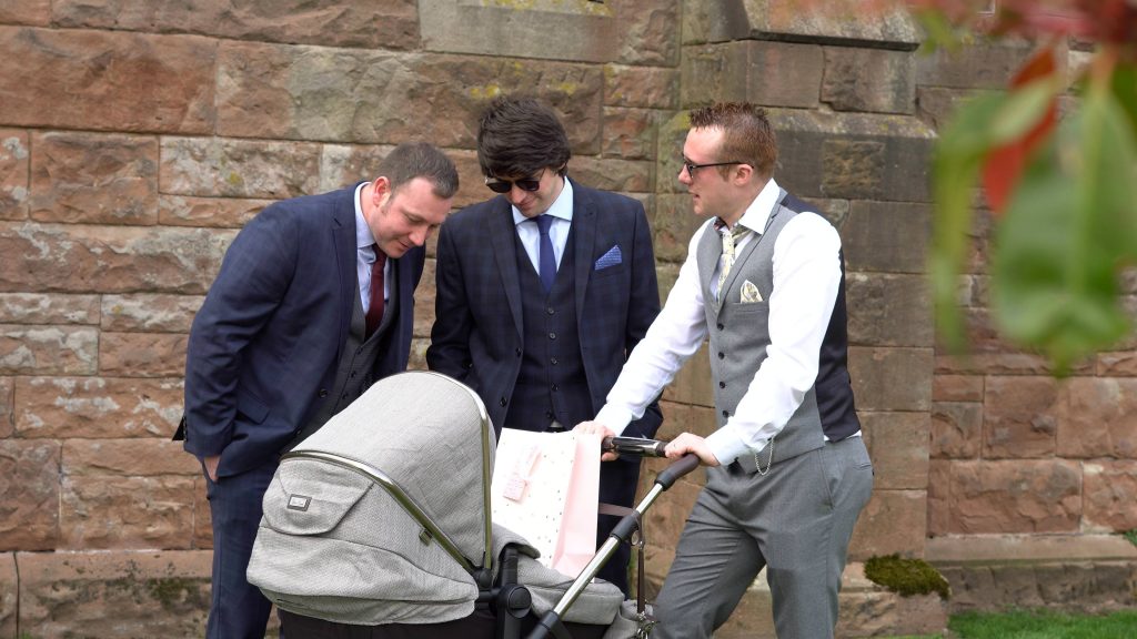 3 guys stand around a pram looking at a baby during the drinks reception at Peckforton castle
