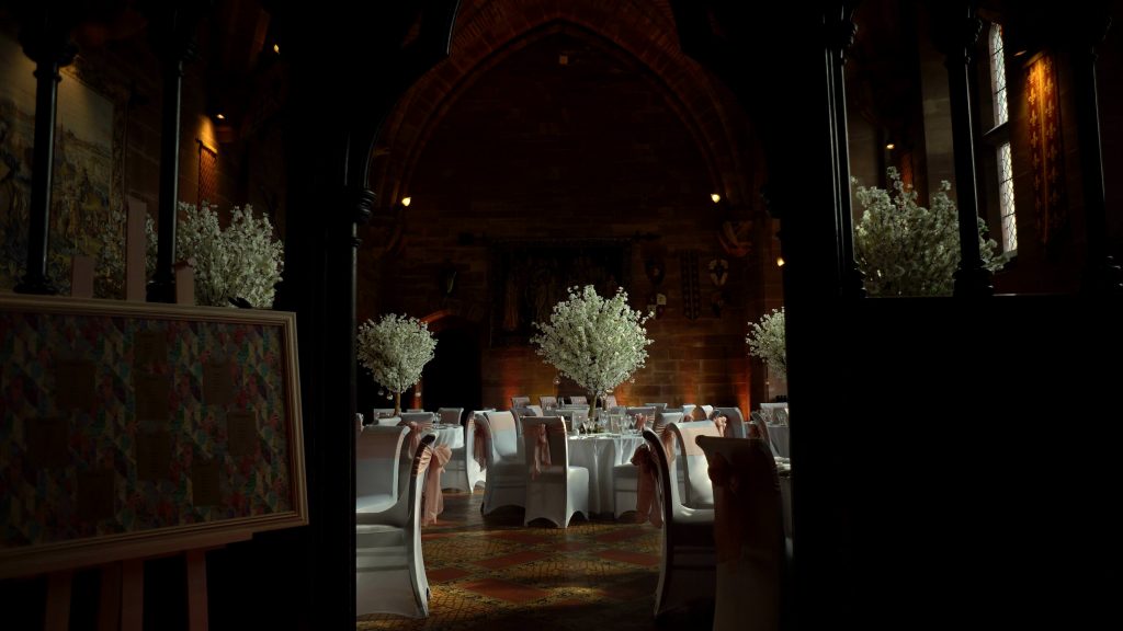 a wide shot from the wedding video of Peckforton castles grand hall ready for a fairytale reception with blossom treets and candles