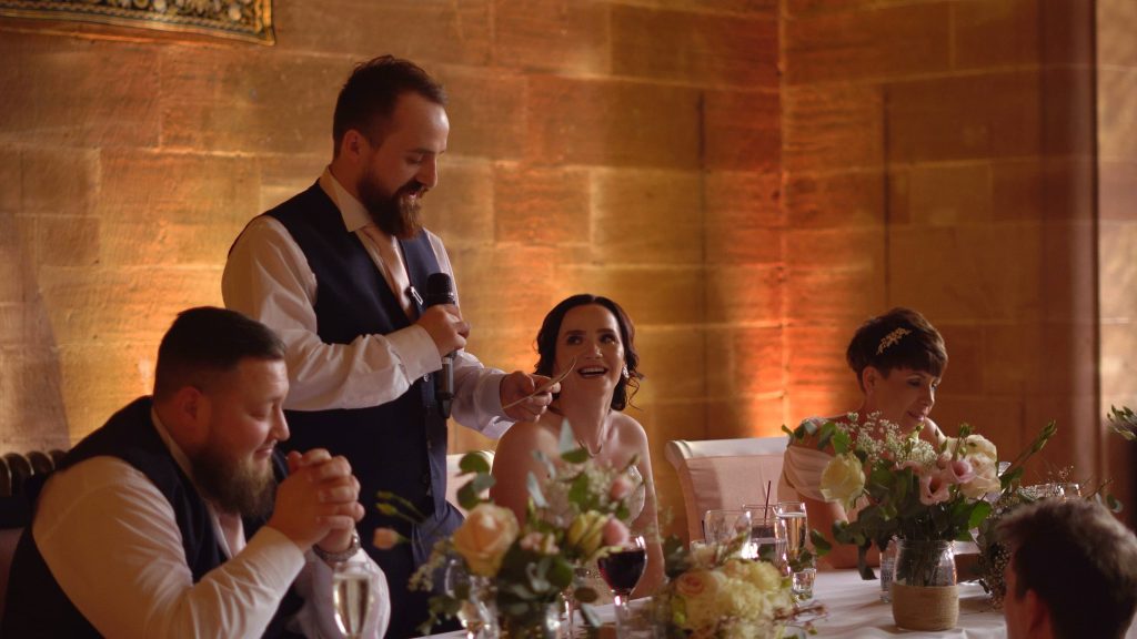 a bride looks up at her groom with a smile as he delivers his grooms speech in the great hall at peckforton castle