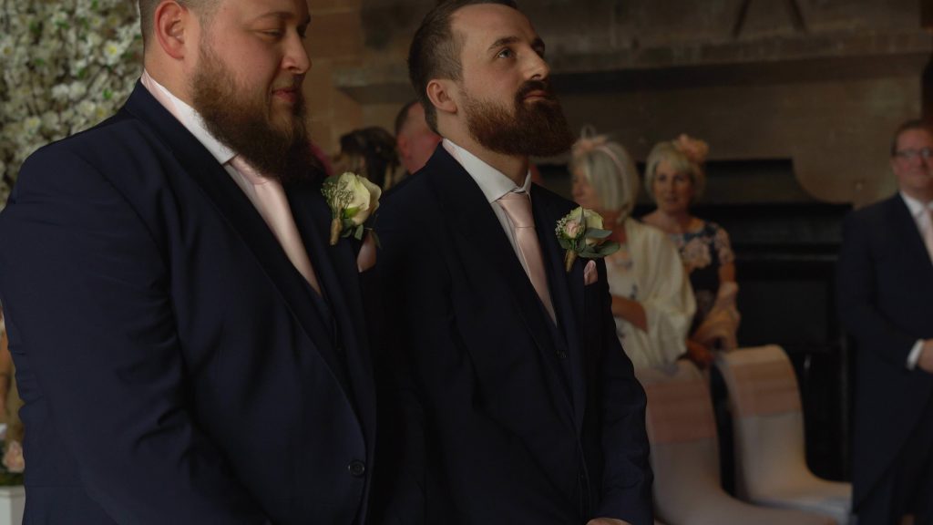 a groom looks up nervously as the guests stand during the wedding ceremony in the great hall at peckforton castle