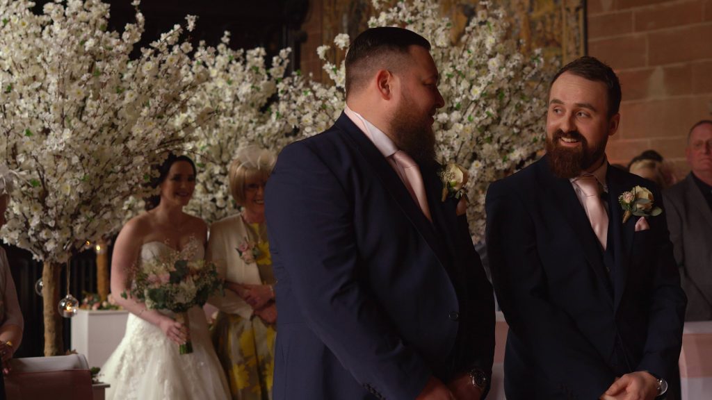 a groom smiles proudly at his best man standing beside him as his bride walks down the aisle during a ceremony at Peckforton castle