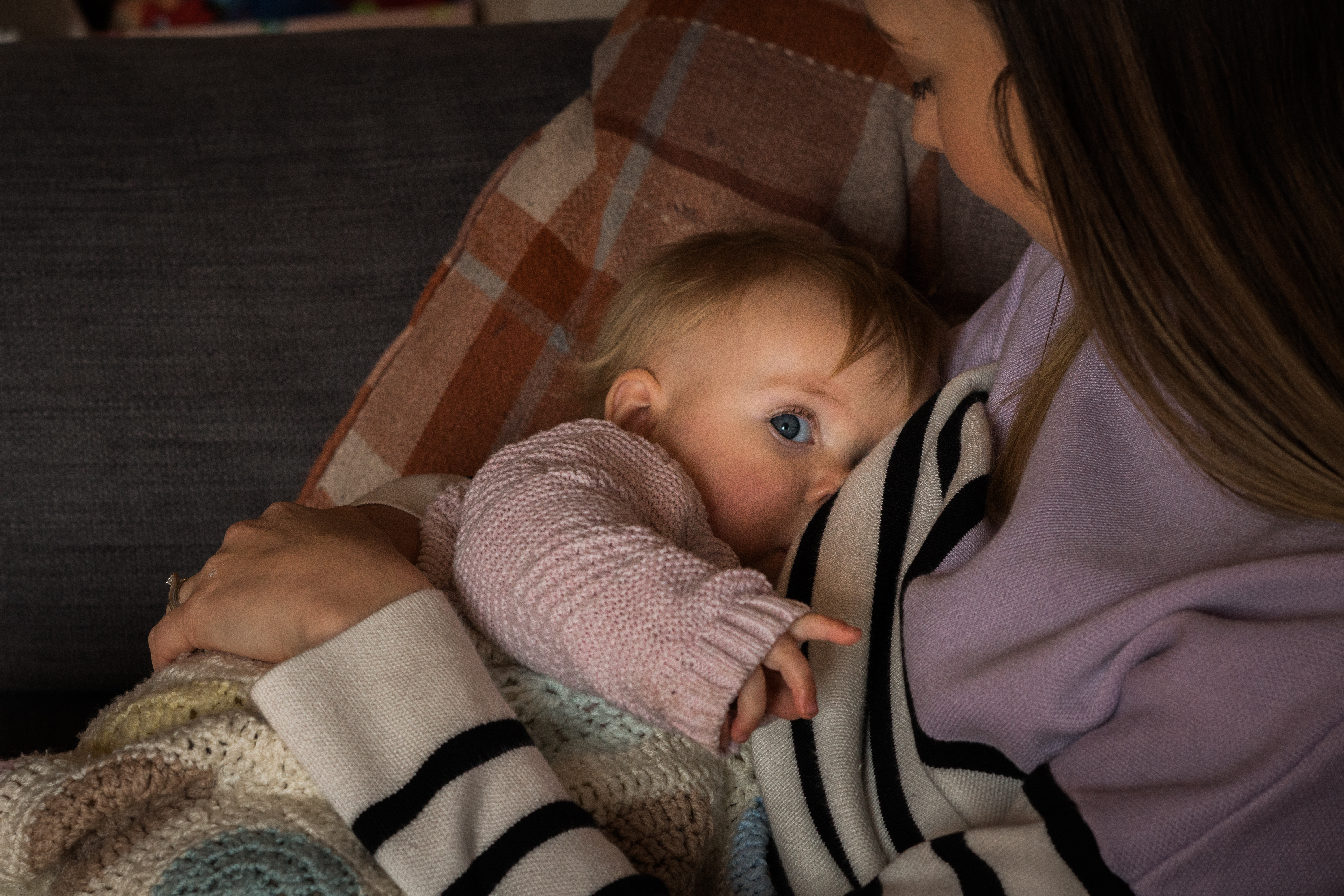 a little girl looks up at the camera whilst breastfeeding during a family photo shoot at home in Lancashire