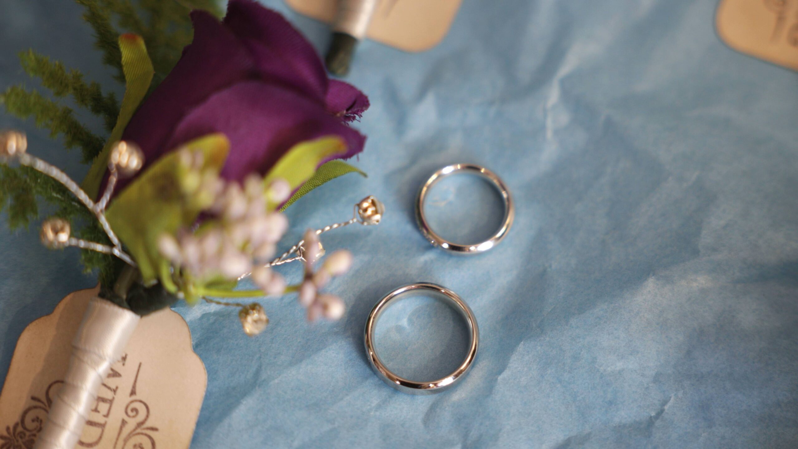 two grooms wedding rings sit by purple button hole