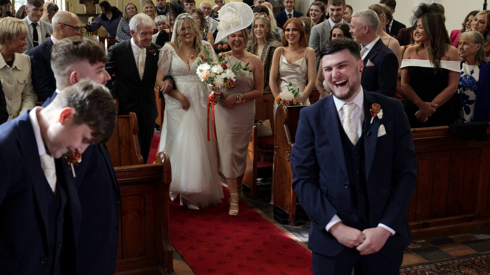 groom laughs at bride and grandad walking down the aisle