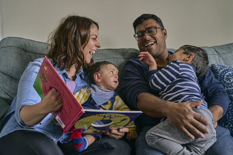 A family sit on the sofa laughing whilst reading at home in Sale