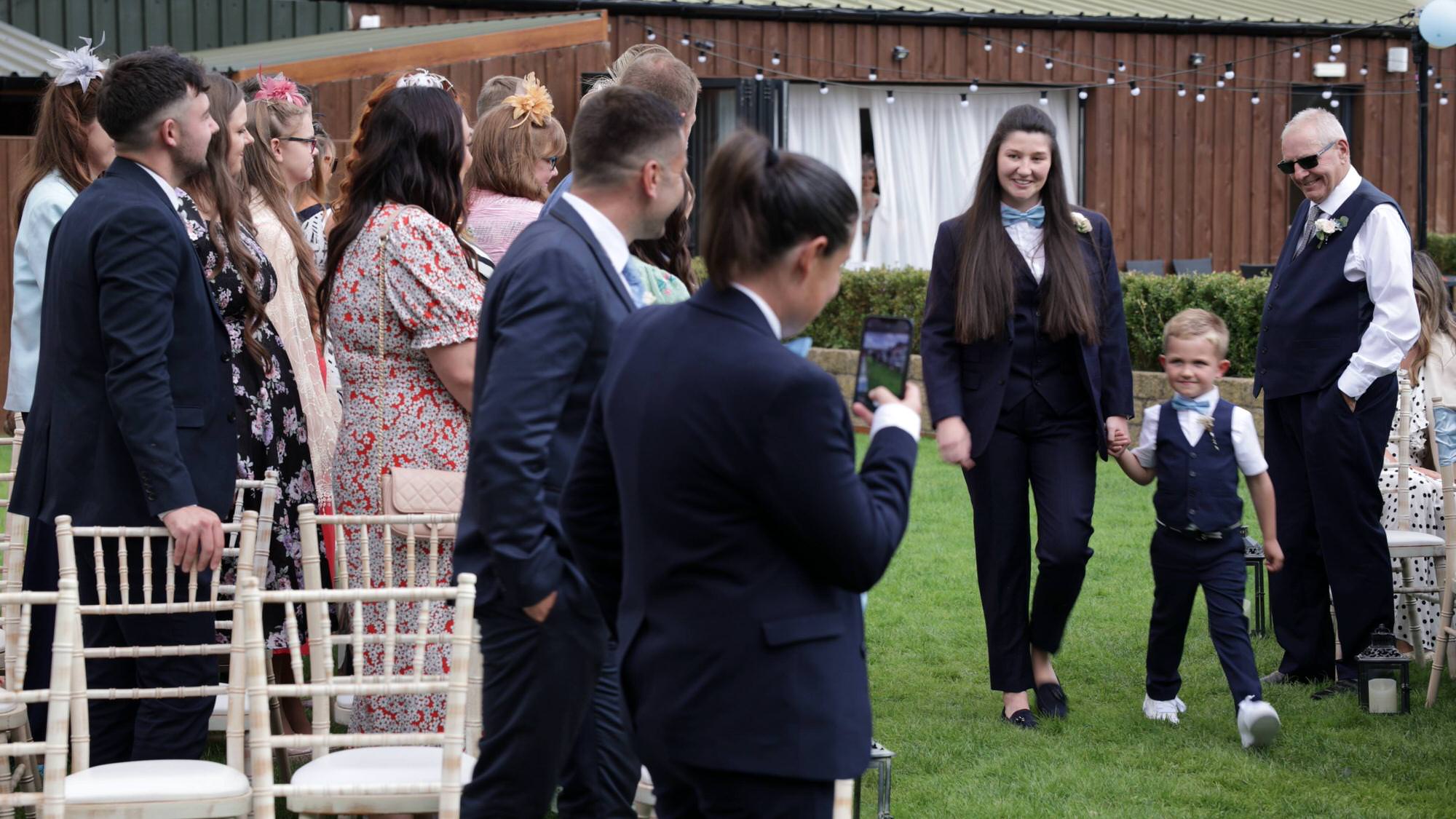 a bride walks down the aisle with a page boy wearing a navy suit