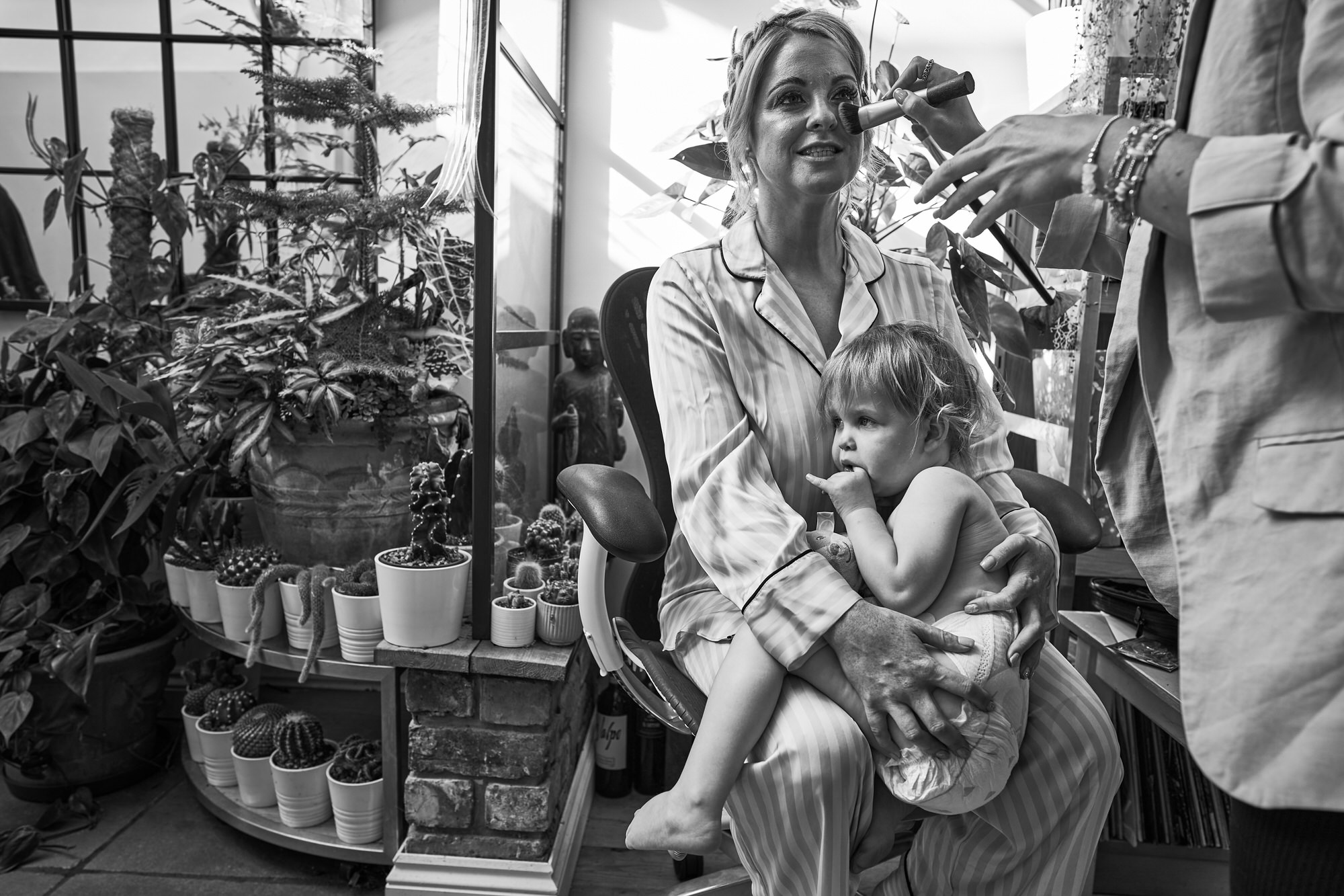 A toddler sits on her Mums knee whilst she has her wedding makeup done