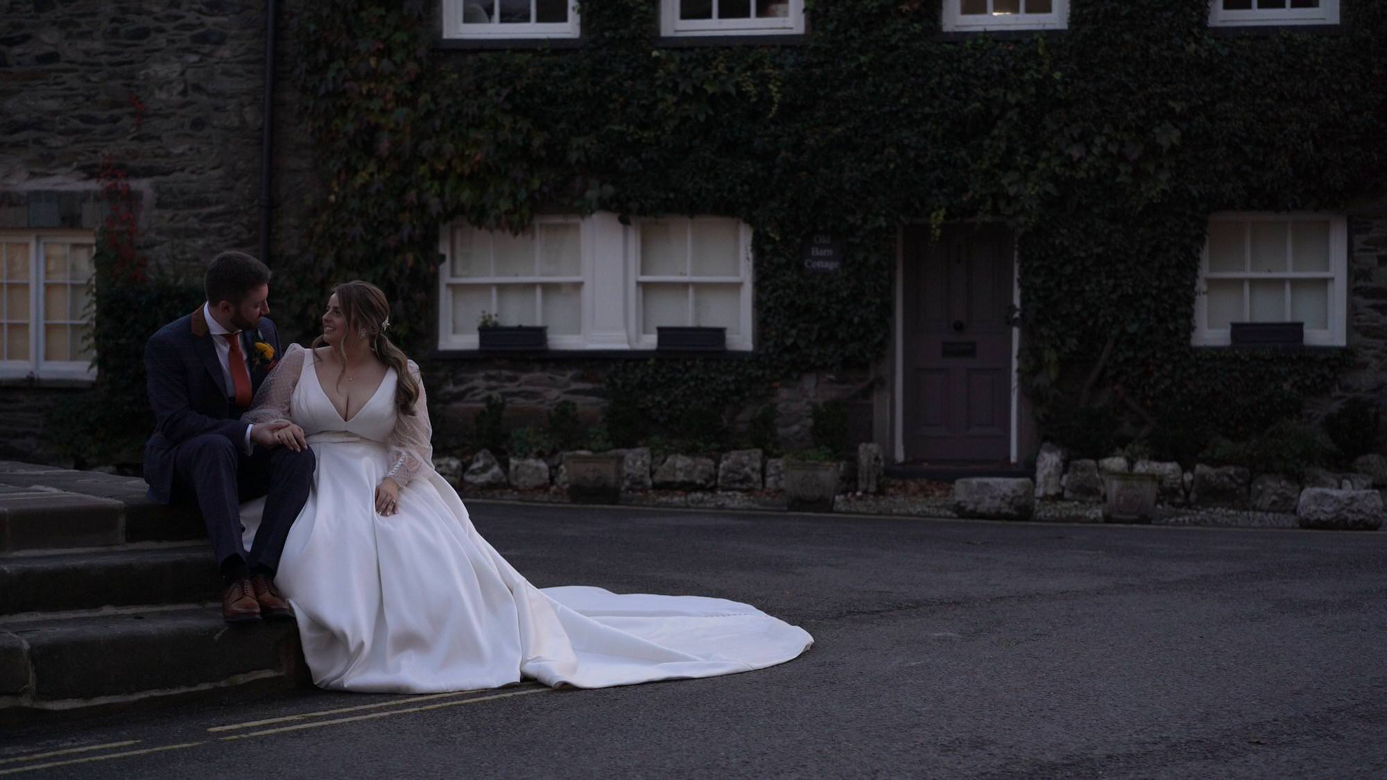 a bride and groom sit on steps outside the famous Cartmel pudding shop