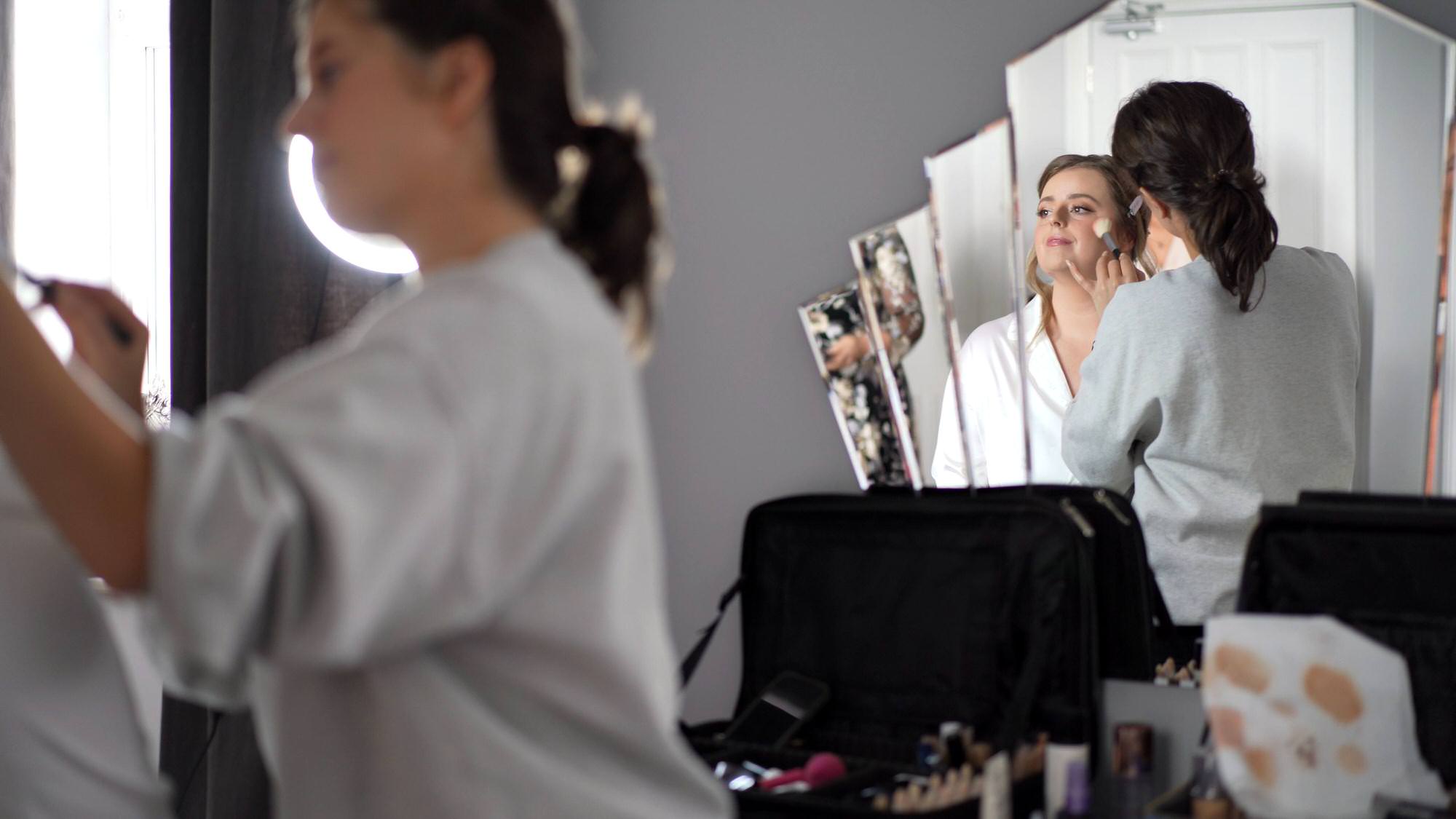 videographer films the bride getting her makeup done in the mirror