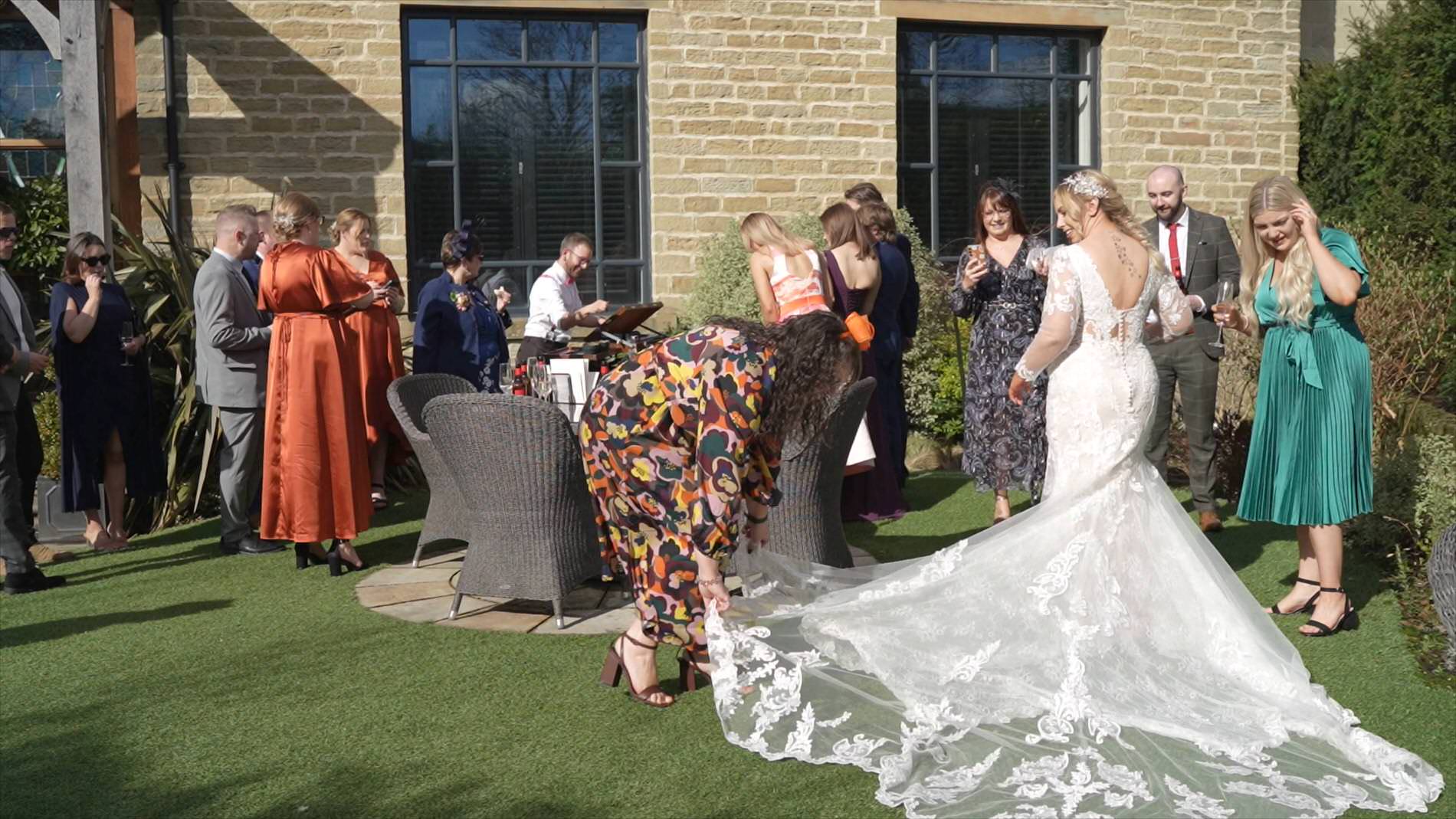 a guest helps lay out the long train for the bride outside Manor House Lindley