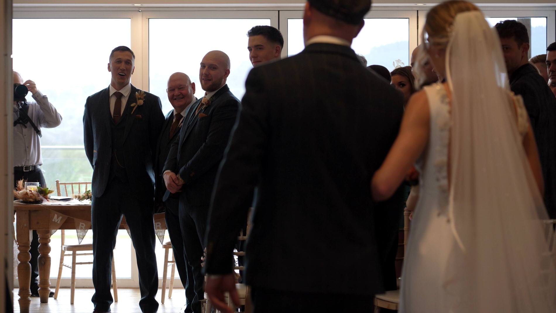 a groom smiles as his bride walks down the aisle at Bashall