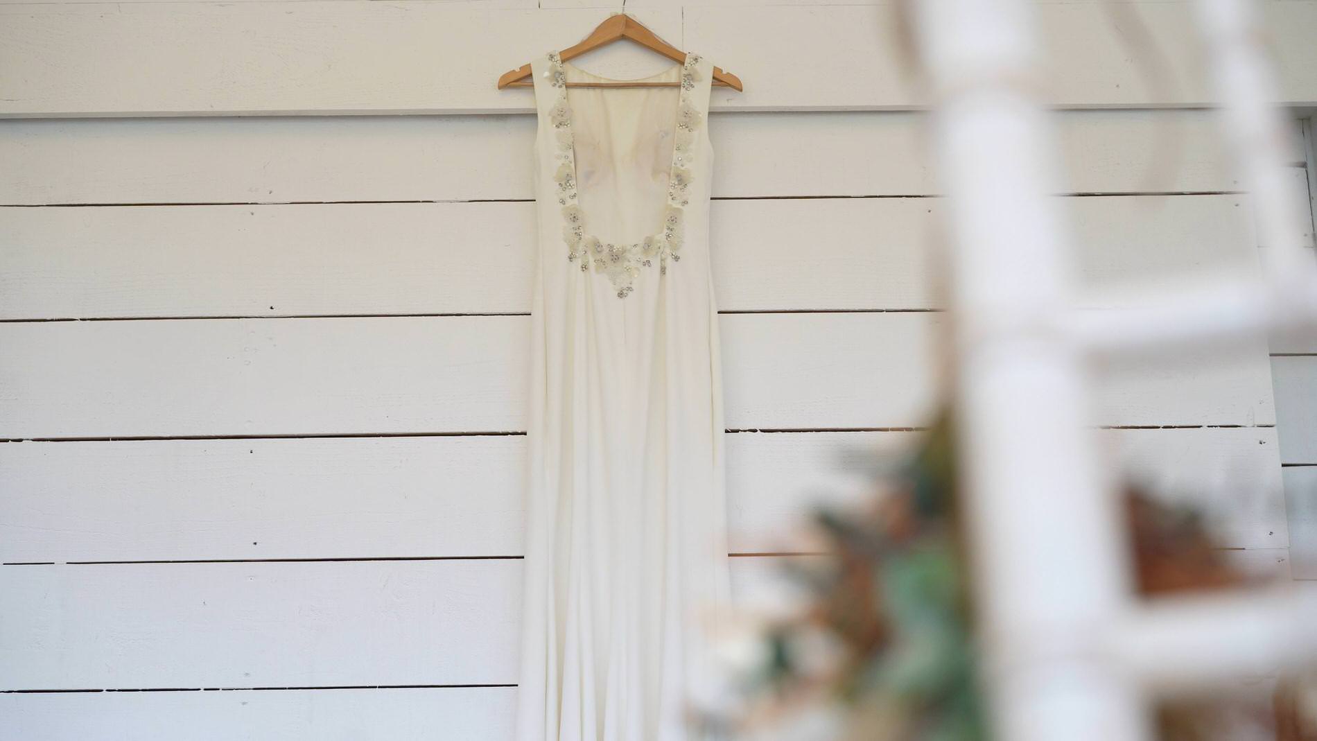 a simple elegant wedding dress hangs on the white wood wall at Bashall