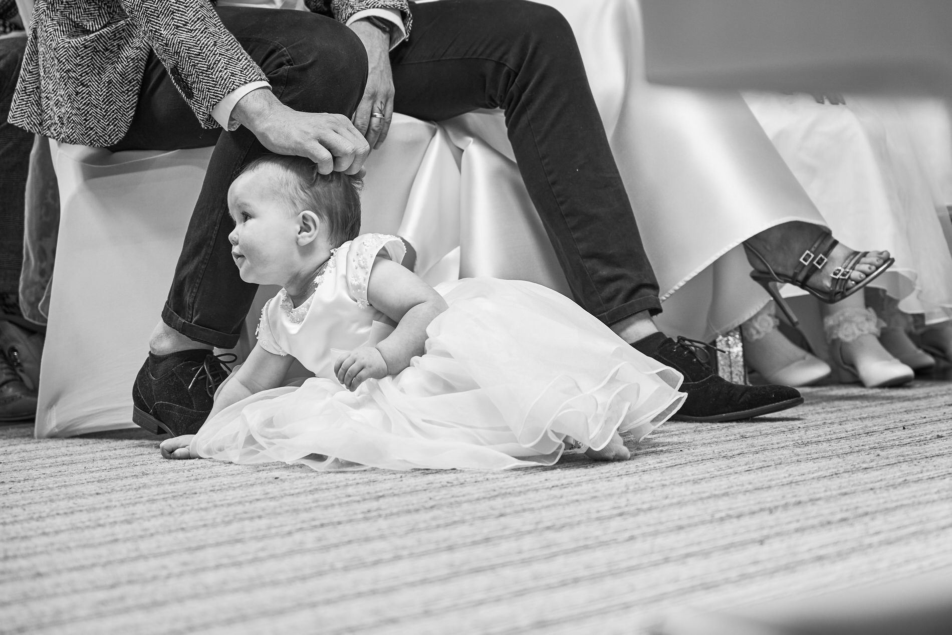 a baby in her christening gown sits on the floor during the ceremony at Holland Hall