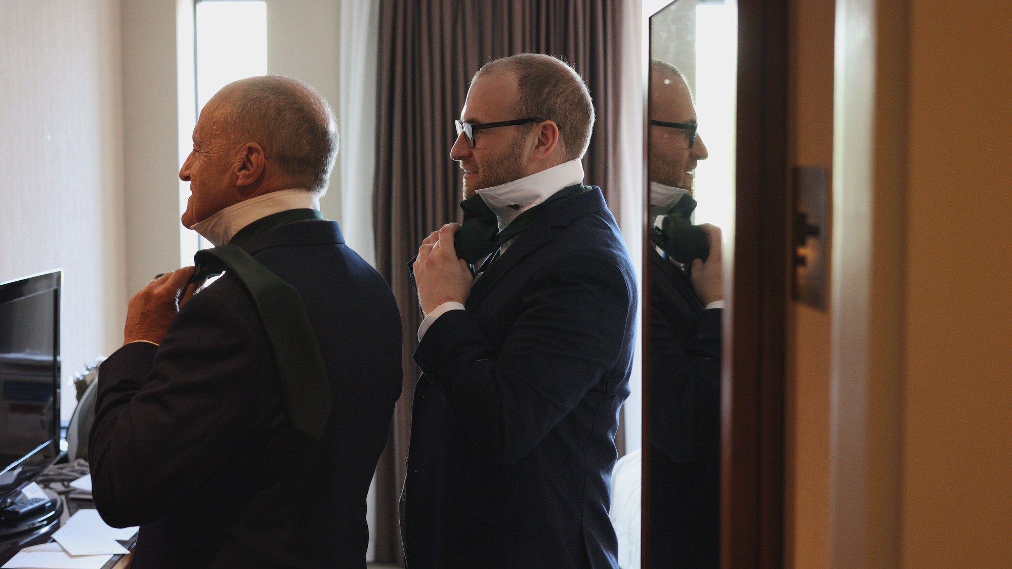 a groom adjusts his tie in the mirror with his Dad